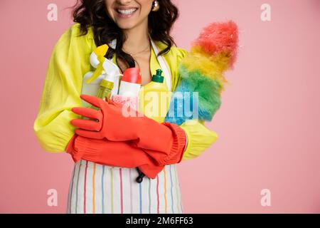 Zugeschnittener Blick auf lächelnde Hausfrau in roten Gummihandschuhen mit vielen Reinigungsmitteln isoliert auf pinkfarbenem Stockbild Stockfoto