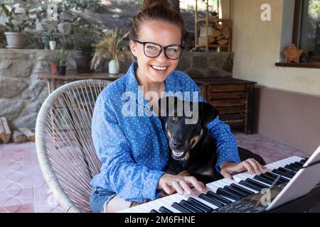 Junge Frau mit Brille, die mit ihrem Hund auf der Terrasse elektrisches Klavier spielt. Stockfoto