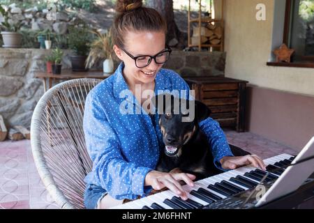 Junge Frau mit Brille, die mit ihrem Hund auf der Terrasse elektrisches Klavier spielt. Stockfoto