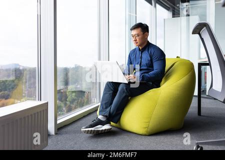 Seriöser und fokussierter Entwickler, Programmierer, der am Arbeitsplatz Code mit einem Laptop schreibt, um Programme und Software zu erstellen, asiatischer Mann in einem modernen Büro in der Nähe von Fenstern. Stockfoto