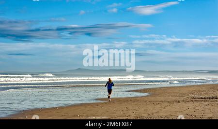 Lossiemouth East Beach Moray Coast Schottland ein blauer Himmel, weiße Wolken und Brandung und ein einsamer Läufer auf dem Sand im Winter Stockfoto