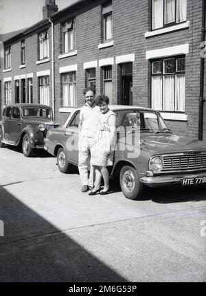 1960er, historisch, Ehemann und Ehefrau stehen neben ihrem Vauxhall Victor Auto der Epoche in einer Straße vor ihrem terrassenförmig angelegten Haus, England, Großbritannien. Ein MG-Auto aus dem letzten Jahrzehnt steht hinter dem Fahrzeug. Stockfoto