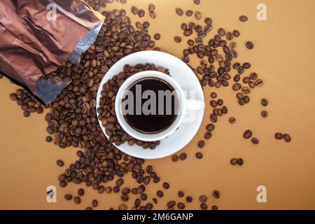 Weiße Tasse schwarzen Americano-Kaffee ohne Milch mit einem Haufen gerösteter Kaffeebohnen, die aus der Papierverpackung verstreut sind. Kaffeebett Stockfoto