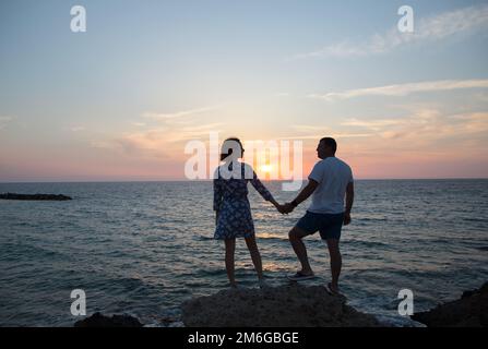 Mann und Frau, er und sie in voller Entwicklung, Silhouetten von verliebten Paaren vor dem Hintergrund des Meeres und des Sonnenuntergangs, halten Händchen. Genießen des Moments, t Stockfoto