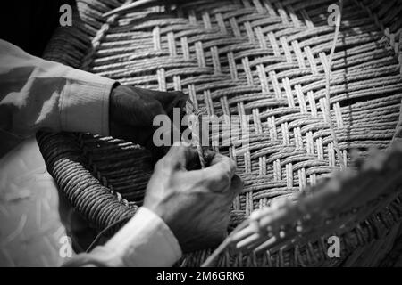 Erfahrener Handwerker, der mit seinen Fingern und Werkzeugen manuell einen detaillierten Bambusholzsessel arbeitet Stockfoto