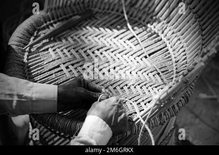 Erfahrener Handwerker, der mit seinen Fingern und Werkzeugen manuell einen detaillierten Bambusholzsessel arbeitet Stockfoto