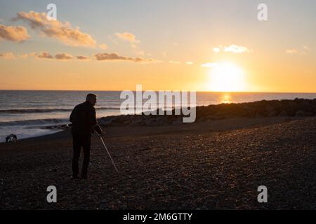 Ein Mann mit einer Krücke am Strand bei Sonnenuntergang in der Küstenstadt Shoreham in Sussex. Foto: Montag, 2. Januar 2023. Foto: Richard Gray/Alamy Stockfoto