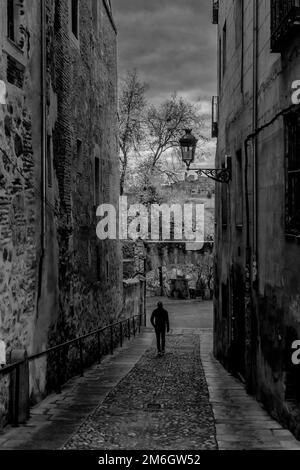 Ein schwarz-weißer Blick auf einen Mann, der eine Gasse in Segovia entlang geht, mit dem ersten Morgengrauen in der Ferne. Stockfoto