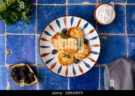 Hüttenkäse-Pfannkuchen auf einem Teller, Brombeeren, Sauerrahm. br Stockfoto