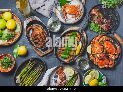 Tisch mit Meeresfrüchtegerichten - gekochte Krabben, Tigergarnelen, gegrillte Tintenfische und Tintenfische auf gusseisernen gegrillten Pfannen und Tellern, Weißwein. Getöntes Foto. Stockfoto