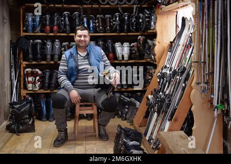 Porträt des leiblichen Eigentümers eines kleinen Skiverleihdienstes in einem Innenraum voller Skier, Stiefel, Helme und anderer Wintersportausrüstung, die vermietet oder verkauft werden kann Stockfoto