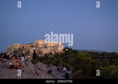 Die Akropolis von Athen, Athen, Griechenland, in der Abenddämmerung, vom Areopagus-Hügel aus gesehen, mit Touristen, die auf dem Hügel im Vordergrund sitzen. Stockfoto