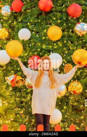 Lächelndes Mädchen in Strickpullover mit riesigen weihnachtsbällen, während es neben einem riesigen glitzernden weihnachtsbaum steht Stockfoto