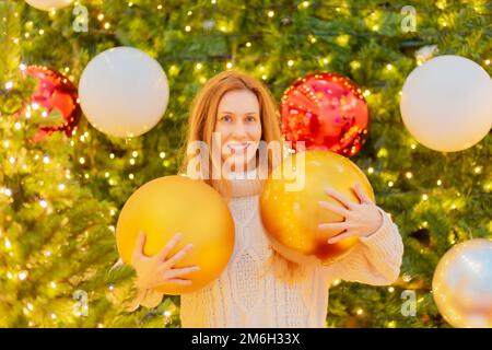 Lächelndes Mädchen mit riesigen Weihnachtsbällen auf dem Hintergrund eines eleganten Weihnachtsbaums. Festliches Poster. Herzlichen Glückwunsch Stockfoto