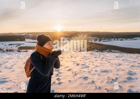 Eine Frau in einem Mantel in einem Schal bläst Schnee von ihren Händen. Stockfoto