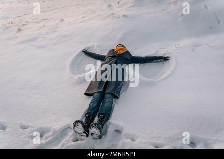 Eine Frau mit Mantel, Schal und Stiefeln liegt auf einem schneebedeckten Feld und macht einen Schneeengel. Stockfoto