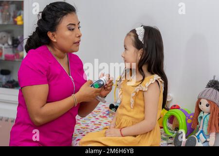 Eine Latina-Kinderärztin, die mit einem Thermometer die Temperatur eines Mädchens messen will, das auf dem Tisch in ihrem Büro sitzt. Stockfoto