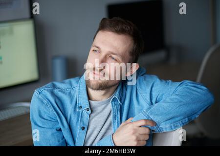 Positive Emotionen mit Computermonitor im Hintergrund. Junger, gutaussehender Mann in Jeansshirt, der sich nach vorne freut. Fröhlicher, bärtiger Mann, der im Heimbüro sitzt. Hochwertige Bilder Stockfoto