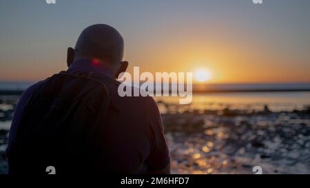 Ein Rückblick auf einen Mann mit Rucksack, der den Blick auf den Sonnenuntergang bewundert Stockfoto