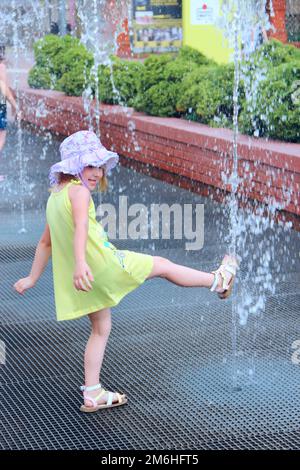 Kleines Mädchen, das versucht, Wasser zu Fuß in den Brunnen der Stadt zu berühren. Die Leute ruhen sich im Stadtpark aus Stockfoto