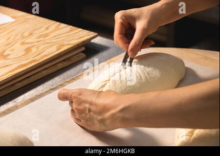 Nahaufnahme der weiblichen Hände einer Bäckerin, die einen Laib Teig mit einer Klinge in einen Laib Brot schneidet, bevor sie im Ofen backen. Craft cr Stockfoto