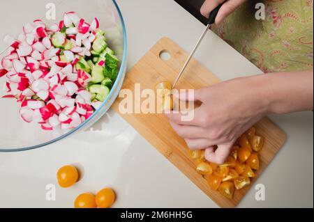 Nahaufnahme von weiblichen Händen, die Kirschtomaten für einen veganen Salat schneiden. Hausmannskost gesunde und gesunde Lebensmittel Stockfoto