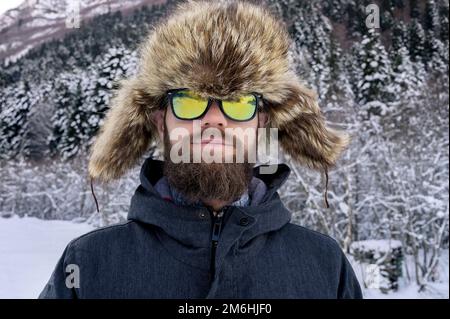 Hübscher, weißer, freundlicher Mann in warmer Winterjacke, Pelzmütze und Sonnenbrille. Ich schaue in die Kamera und lächelte im Winte Stockfoto