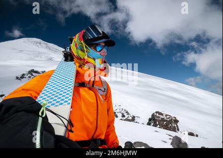 Ein junger, professioneller Skifahrer mit Weitwinkelschuss hält seine Skier, während er hoch in den Bergen steht und auf den schneebedeckten Teil schaut Stockfoto
