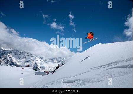 Professioneller Sportler junger männlicher Skifahrer in einem orangefarbenen Skianzug fliegt über die Berge, nachdem er von schneebedeckten Vorsprüngen gesprungen ist. Fre Stockfoto