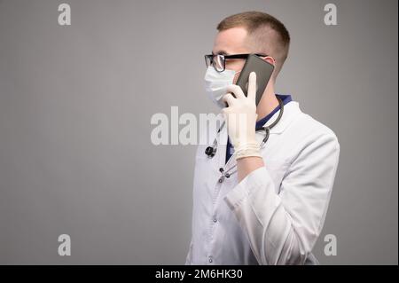 Ein junger weißer Chirurg mit Brille spricht am Telefon, berät sich in einem weißen OP-Kittel und einer Maske auf einem hellen Hintergrund. Mittel Stockfoto