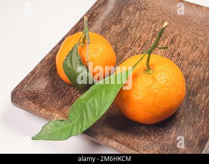 Zwei frische reife Mandarinen mit Blättern und Ästen auf einem dunklen quadratischen Holzteller, Nahaufnahme Stockfoto