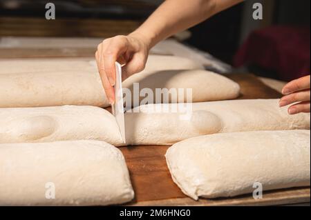 Nahaufnahme der weiblichen Hände einer Bäckerin, die einen Laib Teig mit einer Klinge in einen Laib Brot schneidet, bevor sie im Ofen backen. Craft cr Stockfoto