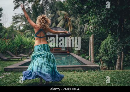 Eine junge Frau in einem blauen Kleid mit blonden Haaren, die in einem tropischen Garten am Pool tanzt Stockfoto