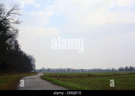 Asphaltpfad, der an einem bewölkten Tag in der italienischen Landschaft von einem bebauten Feld und einem Wald neben einem Wasserstrom umgeben ist Stockfoto