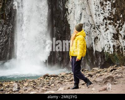 Eine Frau in einer gelben Jacke sieht einen stürmischen, eisigen Wasserfall, der an einem kalten Novembertag nasse Felsen hinunterfließt. Stockfoto