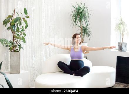 Fröhliche junge schwangere Sportlerin, die auf dem Sofa in einem hellen Raum im Lotus sitzt und ihre Yoga-Session macht Stockfoto