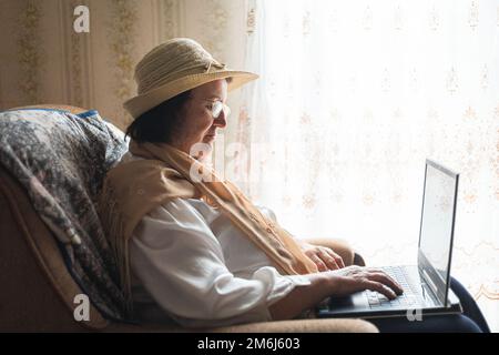 Ältere Frau, die an einem Laptop arbeitet, in einem Stuhl am Fenster sitzt Stockfoto