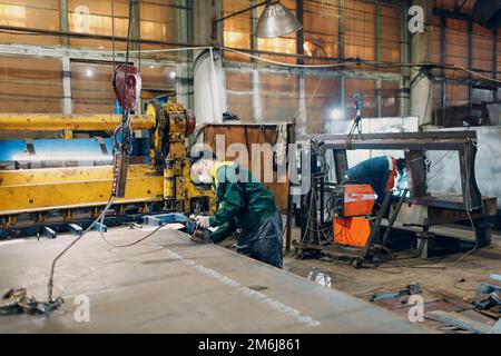 Arbeiter heben Metallblech mit Krankettenzug mit Fernbedienung und Haken in der industriellen Fabrik. Stockfoto