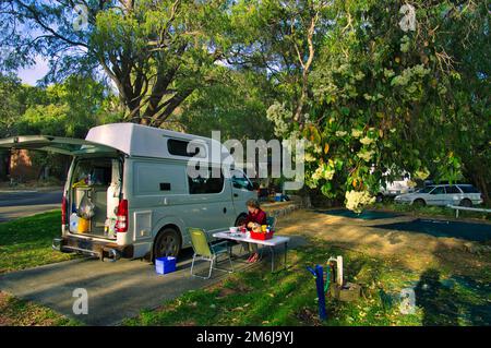 Frau mittleren Alters, die neben einem kleinen Wohnmobil auf einem Camping mit reifen Bäumen in Westaustralien sitzt Stockfoto