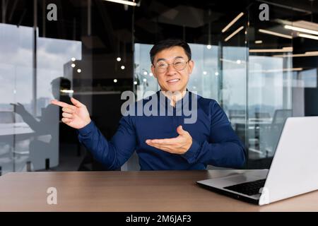 Porträt eines Geschäftsmanns mit Laptop im Büro, lächelnder Mann, der mit Händen und Fingern auf die Seite zeigt, erfolgreicher asiatischer Investor in Hemd und Brille. Stockfoto