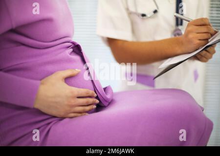 Wunderschöne junge gemischte Rasse weibliche Ärztin mit schwangerer Patientin, die vor dem Teilnehmer sitzt, schriftliche Verschreibung auf dem Formular. Stockfoto