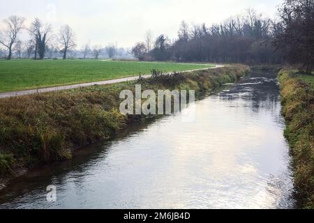 Asphaltierter Pfad mit einem Wald und einem Wasserstrahl am Rande inmitten von bebauten Feldern an einem bewölkten Tag in der italienischen Landschaft Stockfoto