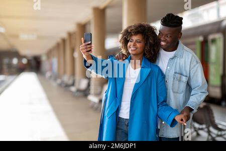 Glückliche Reisende. Ein Junges Schwarzes Paar Mit Smartphone Nimmt Selfie Am Bahnhof Mit Stockfoto