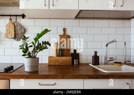 Küchenutensilien aus Messing, Küchenzubehör. Hängeküche mit weißen Fliesen an der Wand und Holztisch. Grüne Pflanze auf Küchenrückseite Stockfoto