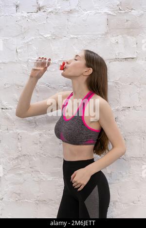 Ein wunderschönes, sportliches Mädchen, das Sportbekleidung trägt, Trinkwasser macht Schnabel nach dem Training, steht neben einer weißen Backsteinwand. Frau af Stockfoto