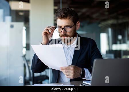Fokussierter Geschäftsmann mittleren Alters, der mit Papieren in modernen Bürogebäuden arbeitet und Finanzdokumente liest Stockfoto