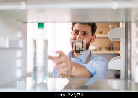 Ein lächelnder weißer Bärtiger in der Schürze öffnet die Tür des leeren Kühlschranks und nimmt eine Flasche Wasser Stockfoto