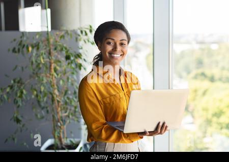 Porträt einer glücklichen Geschäftsfrau, die ihren Laptop im Büro benutzt, neben dem Fenster steht und vor der Kamera lächelt Stockfoto