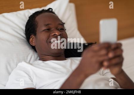 Nahaufnahme Eines Glücklichen Jungen Schwarzen Mannes, Der Während Des Liegens Im Bett Ein Smartphone Benutzt Stockfoto