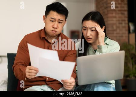 Gestresste asiatische Ehegatten, die Finanzpapiere überprüfen, verärgerter reifer Ehemann und junge Frau, die Kreditpapiere lesen Stockfoto